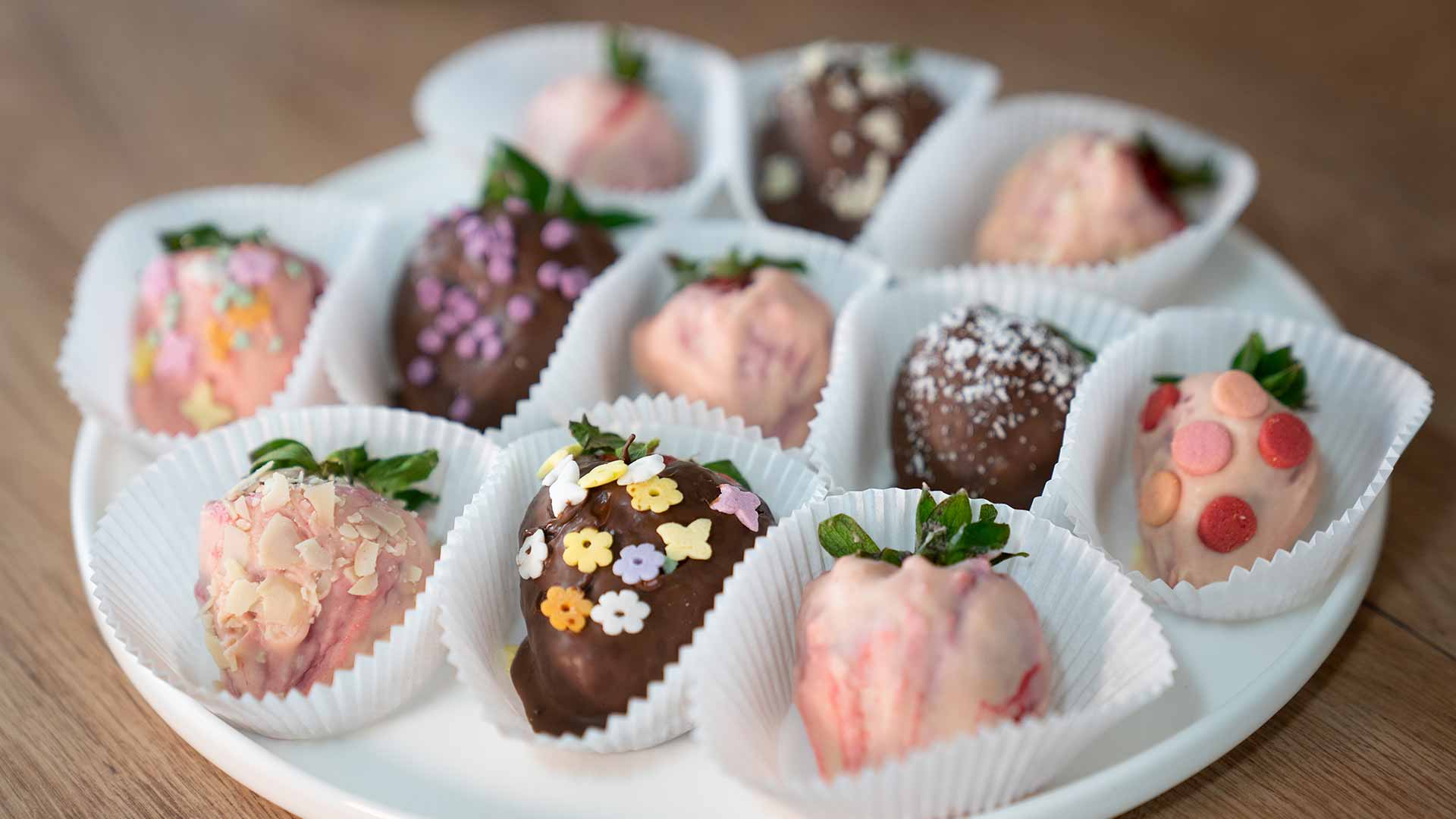 Ein weißer Teller mit schokoladenüberzogenen Erdbeeren, jede mit bunten Streuseln, Nüssen oder Süßigkeiten dekoriert und in weißen Pappbechern platziert.