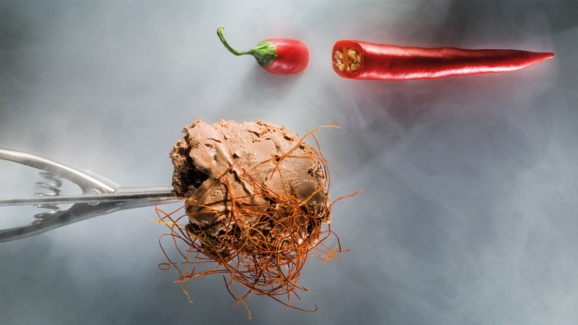 Eine Kugel Schokoladeneis, durchzogen von dünnen Chilifäden, gehalten von einer Metallzange, mit einer ganzen roten Chili und einer geschnittenen Chili auf einem rauchig-grauen Hintergrund.