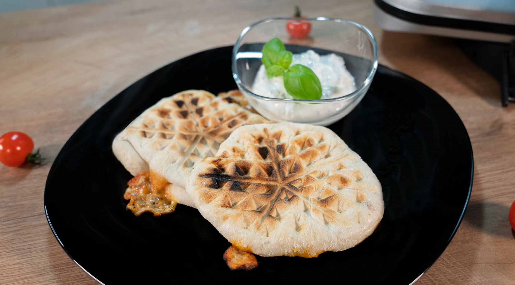 Zwei gegrillte Fladenbrote mit geschmolzenem Käse auf einem schwarzen Teller, serviert mit einer Glasschale cremigem Dip, garniert mit frischem Basilikum. Im Hintergrund liegt eine Kirschtomate auf einer Holzoberfläche.