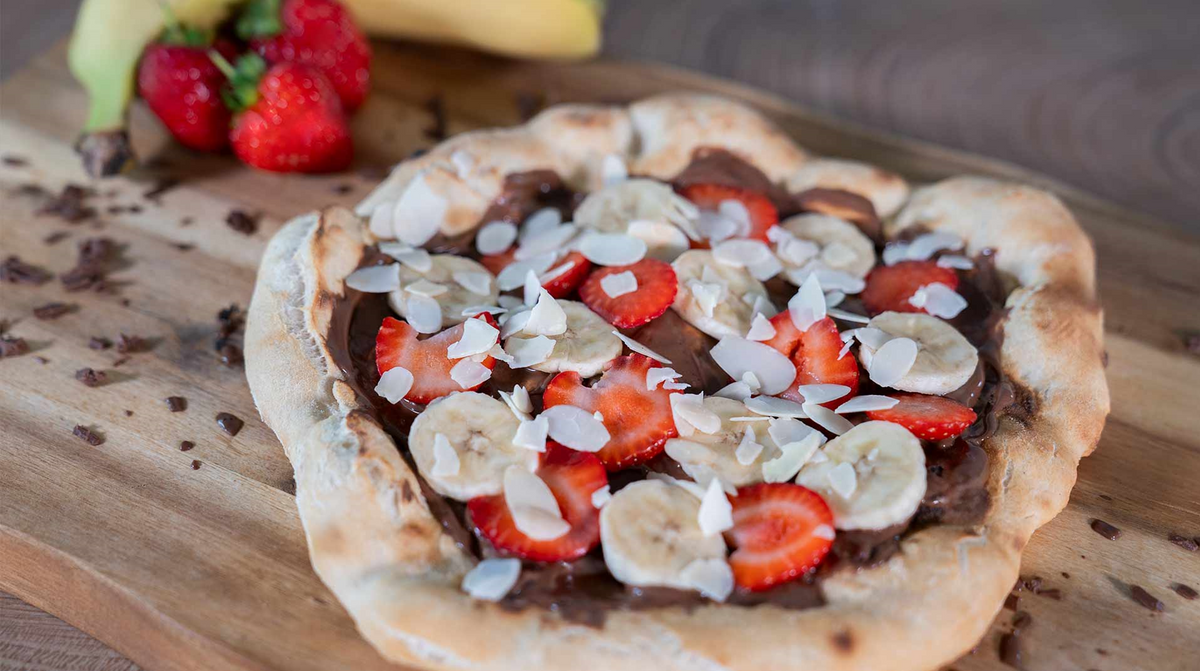 Eine Dessertpizza auf einem Holzbrett, belegt mit Schokoladenaufstrich, Bananenscheiben, Erdbeerscheiben und Mandelblättchen. Im Hintergrund sind frische Erdbeeren und Bananen zu sehen.