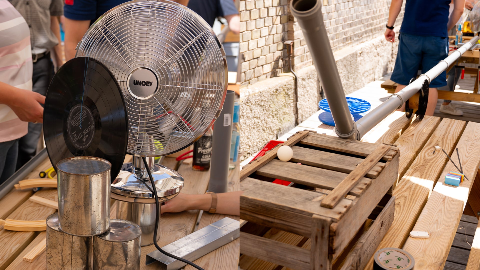 Auf einem Holztisch steht eine selbstgebaute Rube-Goldberg-Maschine mit einem Ventilator, einer Schallplatte, Metalldosen, Rohren, einer Kiste und verschiedenen Gegenständen, während die Leute draußen in der Nähe stehen.