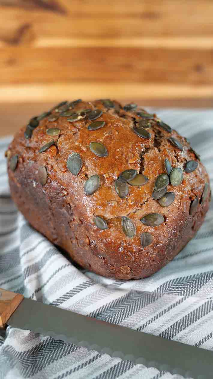 Ein Laib dunkles, rustikales Brot mit Kürbiskernen liegt auf einem gestreiften Tuch, daneben ein Brotmesser. Der Hintergrund ist aus Holz und schafft eine warme, gemütliche Atmosphäre.