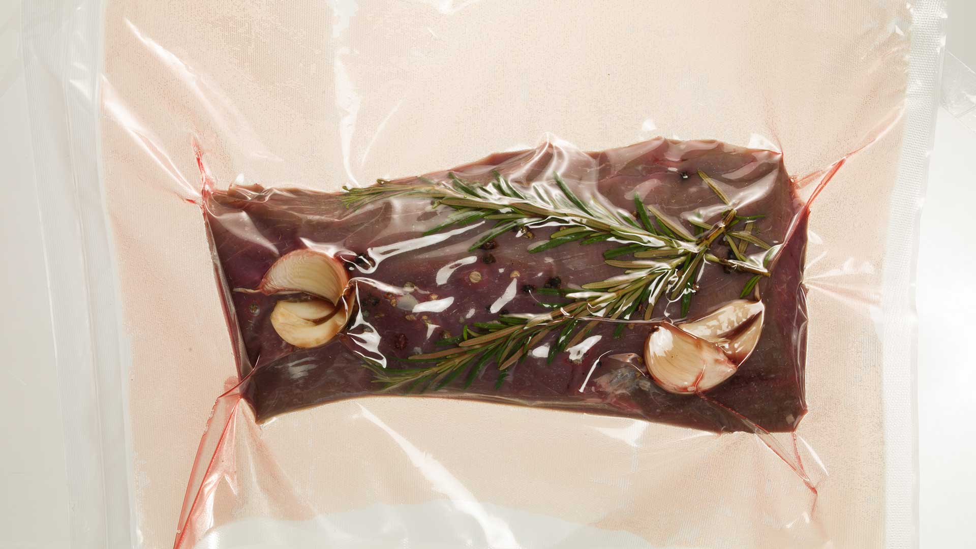 Ein rohes Steak wird mit Rosmarinzweigen, Knoblauchzehen und gemahlenem Pfeffer in einem Vakuumbeutel versiegelt und ist bereit zum Sous-Vide-Garen.