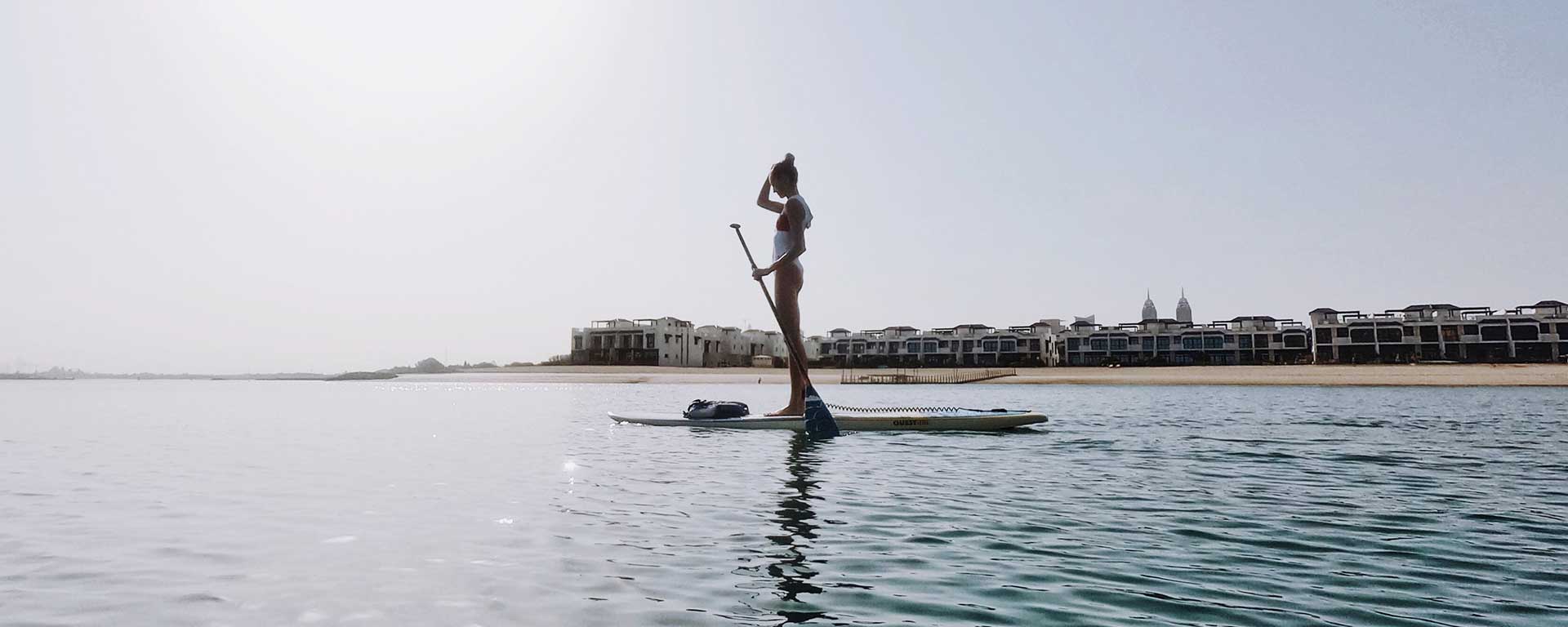 Eine Frau im Badeanzug steht auf einem Paddleboard in ruhigem Wasser in Ufernähe, im Hintergrund sind moderne Gebäude und ein klarer Himmel zu sehen.