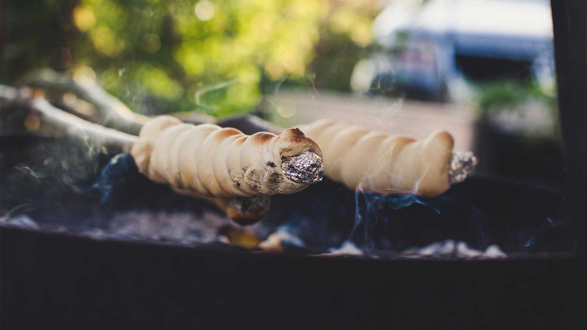 Zwei mit Teig umwickelte Stäbchen werden über einem Grill oder Lagerfeuer geröstet. Rauch steigt auf, und im Hintergrund ist das Grün verschwommen. Die Enden der Stäbchen sind mit Alufolie umwickelt.
