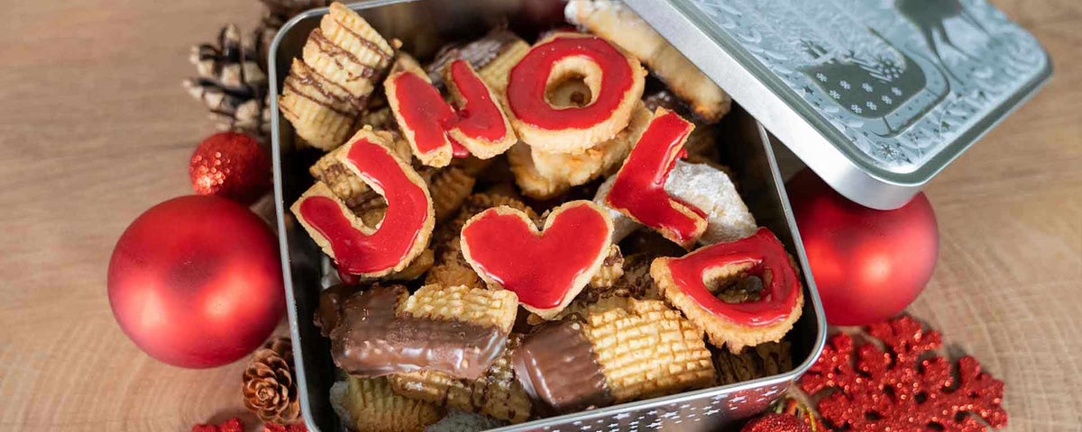 Eine offene Blechdose mit verschiedenen Keksen, einige davon mit rotem Zuckerguss, der „UNO“ und ein Herz buchstabiert. Roter Weihnachtsschmuck und Tannenzapfen liegen rund um die Dose auf einer Holzoberfläche.