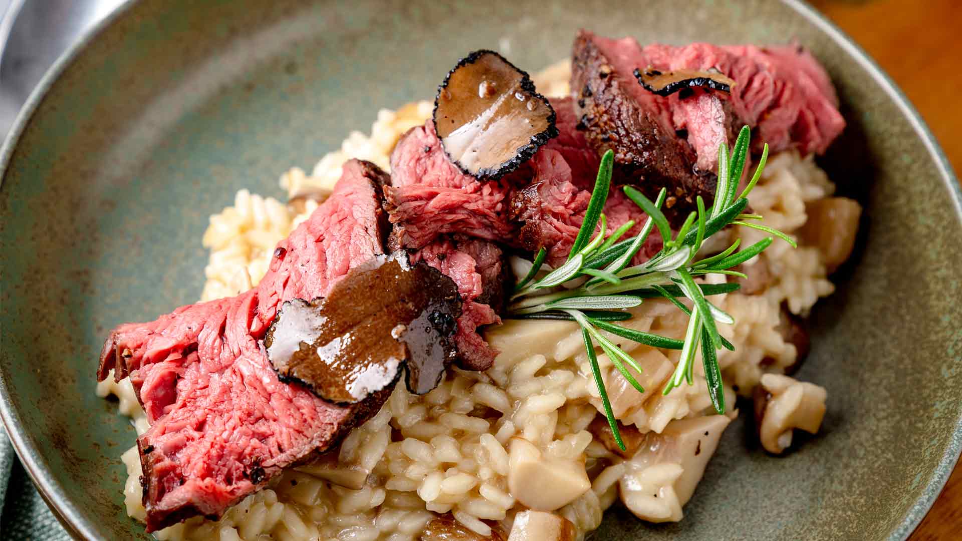 Ein Teller mit cremigem Pilzrisotto, belegt mit medium gebratenen Steakscheiben, dünn geschnittenen schwarzen Trüffeln und einem Zweig frischem Rosmarin.