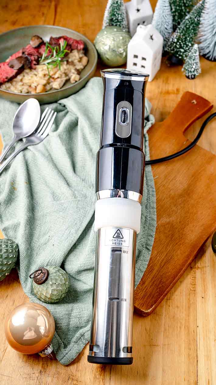 Ein Sous-Vide-Immersionszirkulator steht auf einem hölzernen Schneidebrett neben Weihnachtsdekorationen, Besteck und einer grünen Serviette, im Hintergrund ist ein festlicher Teller mit Essen zu sehen.