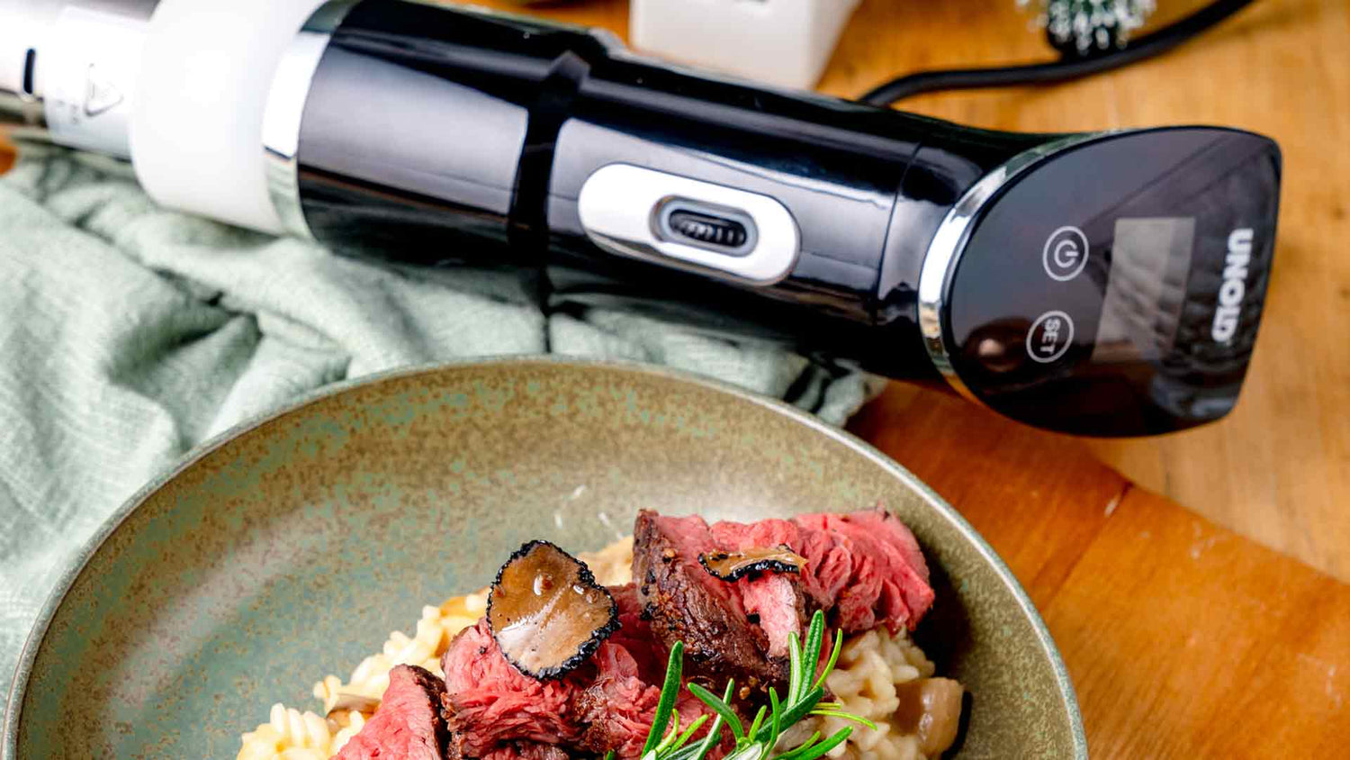 Ein Teller Risotto mit Scheiben von blutigem Rindfleisch, schwarzem Trüffel und Rosmarin steht auf einem Holztisch neben einem modernen Sous-Vide-Tauchzirkulator in Schwarz und Silber.