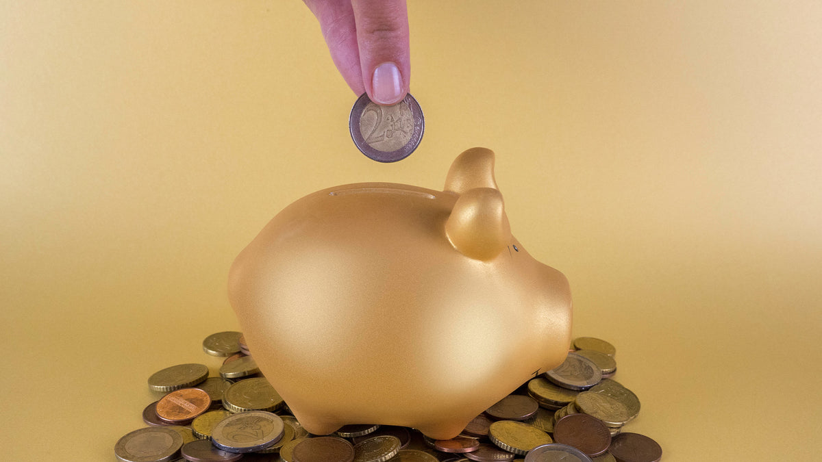 Eine Hand legt eine 2-Euro-Münze in ein goldenes Sparschwein, umgeben von verstreuten Münzen auf gelbem Hintergrund.