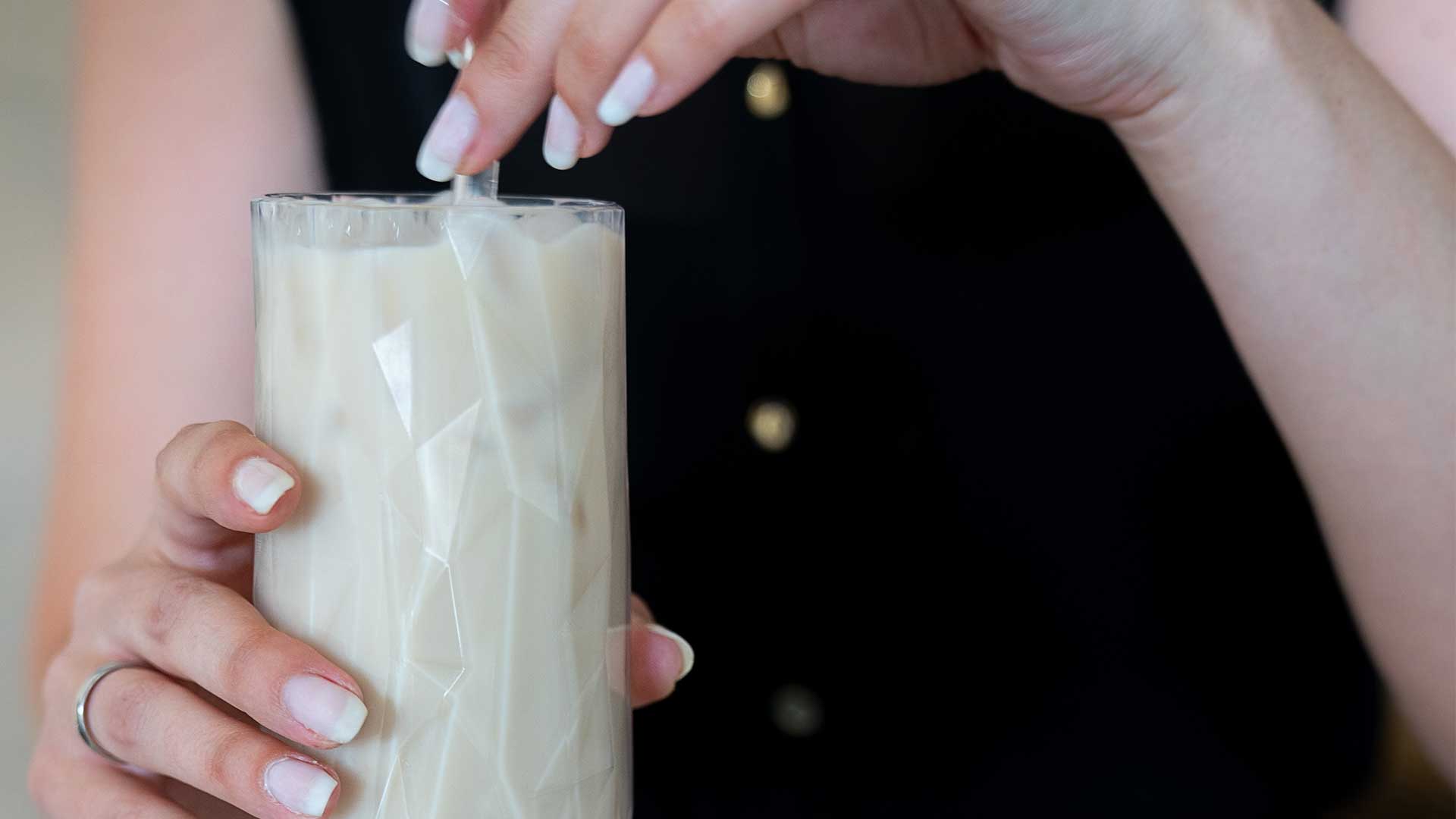 Eine Person mit hellrosa manikürten Nägeln rührt mit einem Strohhalm in einem Glas Eismilch oder einem cremigen Getränk. Das Glas weist ein geometrisches Muster auf und die Person trägt ein schwarzes Oberteil mit goldenen Knöpfen.