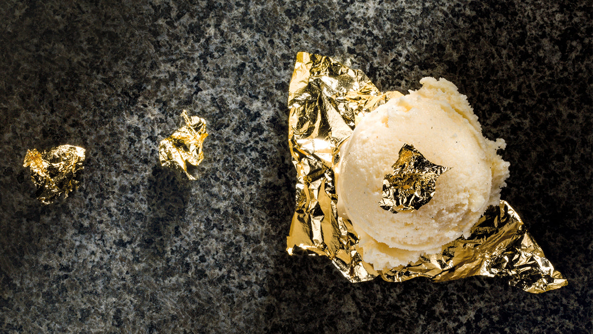 Eine Kugel Vanilleeis wird auf ein Blatt Goldblatt gelegt, mit einem kleinen Stück Goldblatt oben drauf und zwei weiteren Stücken daneben, alles auf einer dunklen, strukturierten Oberfläche.