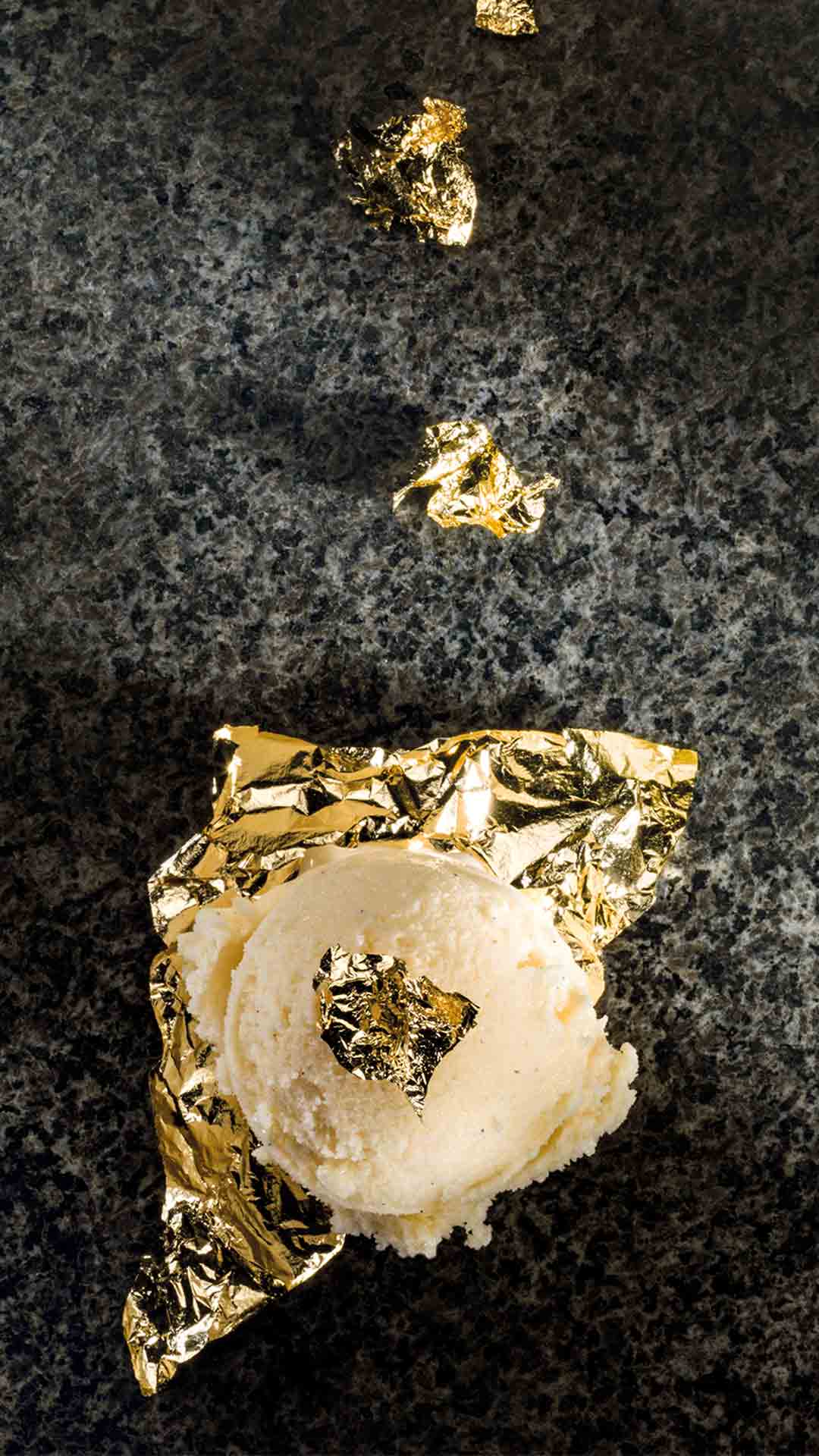 Eine Kugel Vanilleeis liegt auf einer dunklen, strukturierten Oberfläche, teilweise in Goldfolie eingewickelt und darüber verstreut mit kleinen Stücken Blattgold.