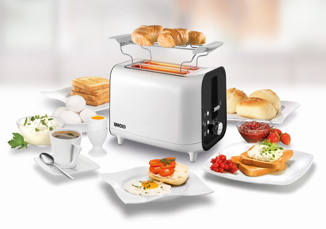 Der Unold Toaster Shine White wird auf einer weißen Oberfläche mit Croissants darauf präsentiert, umgeben von Tellern mit Toast, Eiern, Kaffee, Brötchen, Obst und Aufstrichen vor einem sanft verschwommenen Hintergrund.