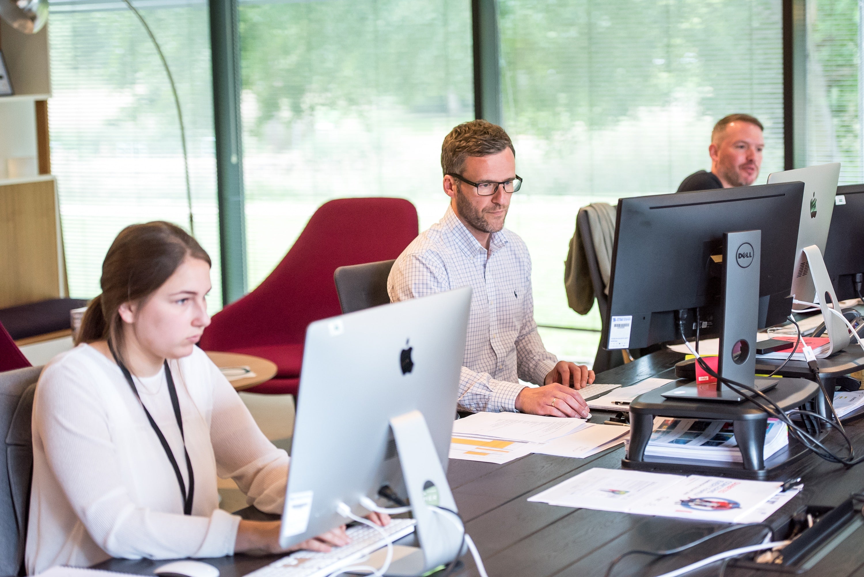 Drei Personen arbeiten an Schreibtischen mit Computern in einem modernen Büroraum mit großen Fenstern, natürlichem Licht und Papieren auf den Schreibtischen.