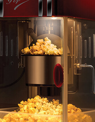 Eine Nahaufnahme der Unold Popcornmaschine Retro im Einsatz. Frisches Popcorn quillt aus dem Metallkessel in das untere Fach. Die Maschine verfügt über ein Klarglasgehäuse und rote Akzente.