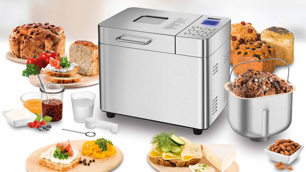 Ein Brotbackautomat aus Edelstahl, umgeben von verschiedenen Brotsorten, belegten Brotscheiben, Marmeladengläsern und kleinen Schälchen mit Butter und Aufstrichen auf einer weißen Fläche.