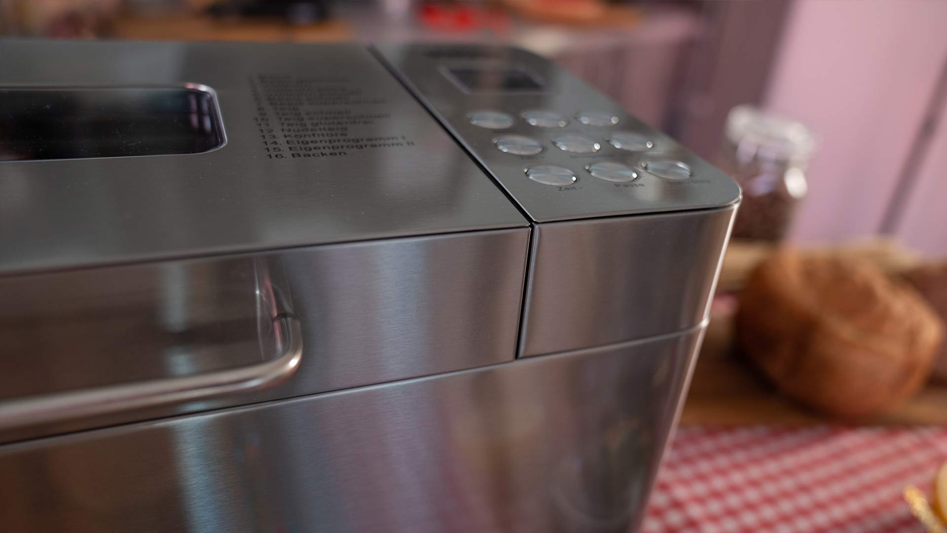 Nahaufnahme eines Brotbackautomaten aus Edelstahl mit Bedientasten und einer Programmliste. Im Hintergrund liegen ein Laib Brot, ein Glas und ein rot kariertes Tuch auf einem Holztisch.
