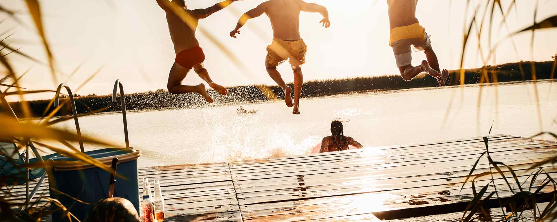 Drei Personen in Badebekleidung springen von einem Holzsteg in einen sonnenbeschienenen See, während eine weitere Person bereits im Wasser liegt. Die Szene ist hell und sommerlich, mit plätscherndem Wasser und warmem Sonnenlicht.