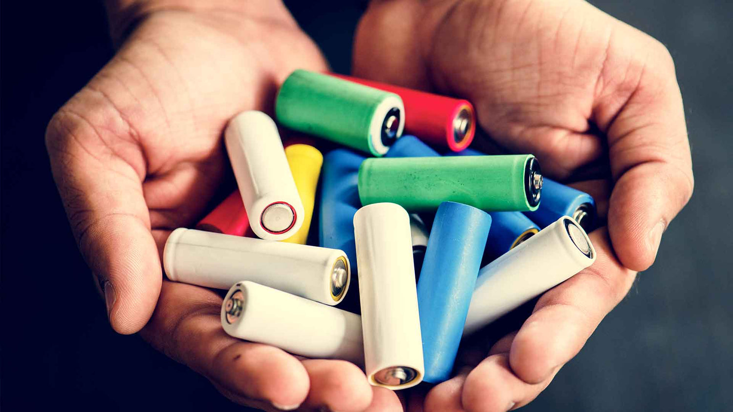Ein Paar Hände, die vor einem dunklen Hintergrund mehrere bunte zylindrische Batterien halten, darunter rote, blaue, grüne, weiße und gelbe.