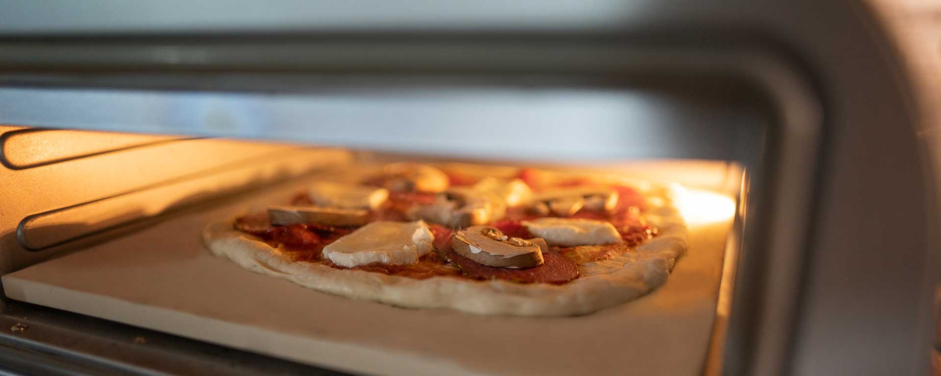 Eine selbstgemachte Pizza mit Käse und Champignons wird auf einem Pizzastein im Ofen gebacken, wobei die Ofentür teilweise geöffnet und der Innenraum warm beleuchtet ist.