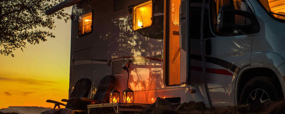 Ein gemütlicher Wohnwagen mit offener Tür und warmen Lichtern im Inneren, der bei Sonnenuntergang im Freien geparkt ist. Draußen unter einem Baum sind zwei Stühle und Laternen aufgestellt.