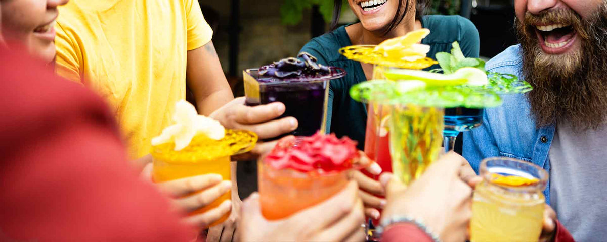 Eine Gruppe von Freunden lächelt und lacht, während sie in fröhlicher Atmosphäre im Freien mit bunten Cocktails anstoßen und gemeinsam ihre Getränke hochhalten.
