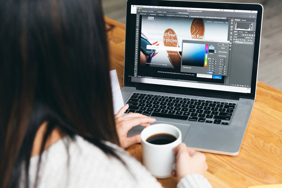 Eine Person mit einer weißen Tasse sitzt an einem Holztisch und arbeitet an einem Laptop, auf dem ein Grafikdesignprogramm mit einer Bildbearbeitungsoberfläche und einem Farbauswahlwerkzeug zu sehen ist.