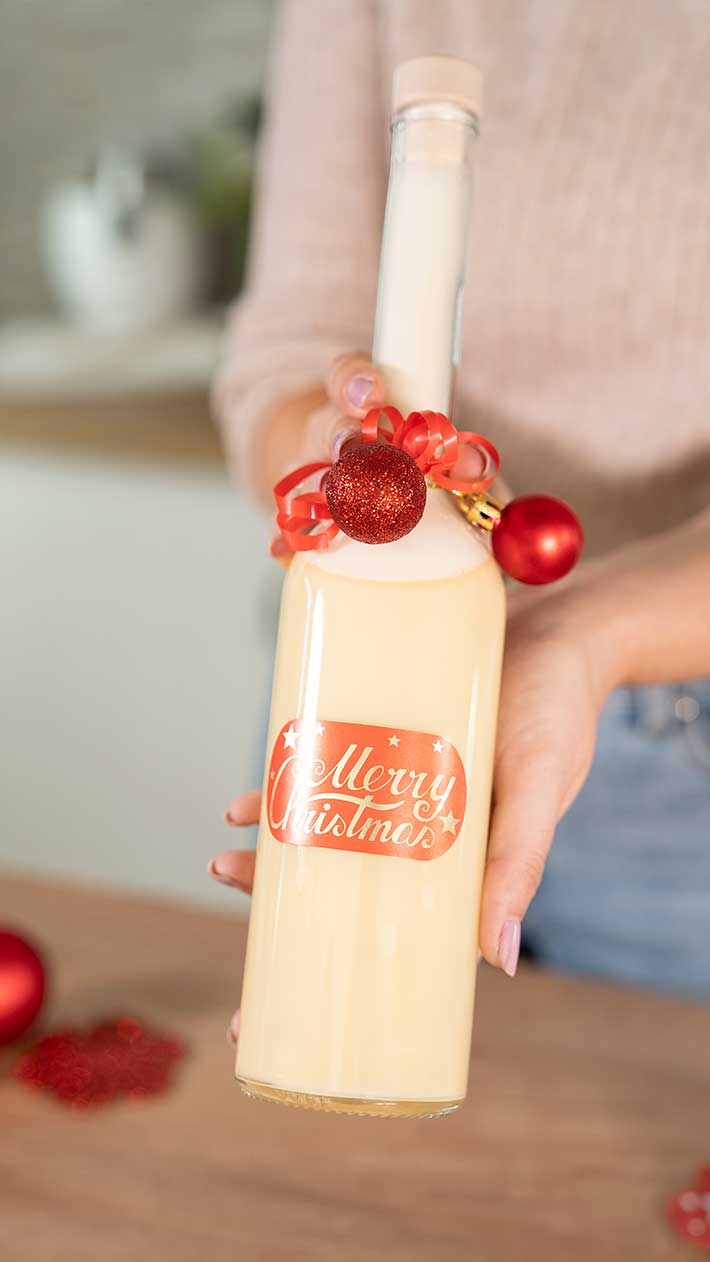 Eine Person hält eine Flasche Sahnelikör mit dem Etikett „Frohe Weihnachten“, verziert mit einer roten Schleife und Weihnachtsschmuck. Der Hintergrund ist leicht verschwommen.