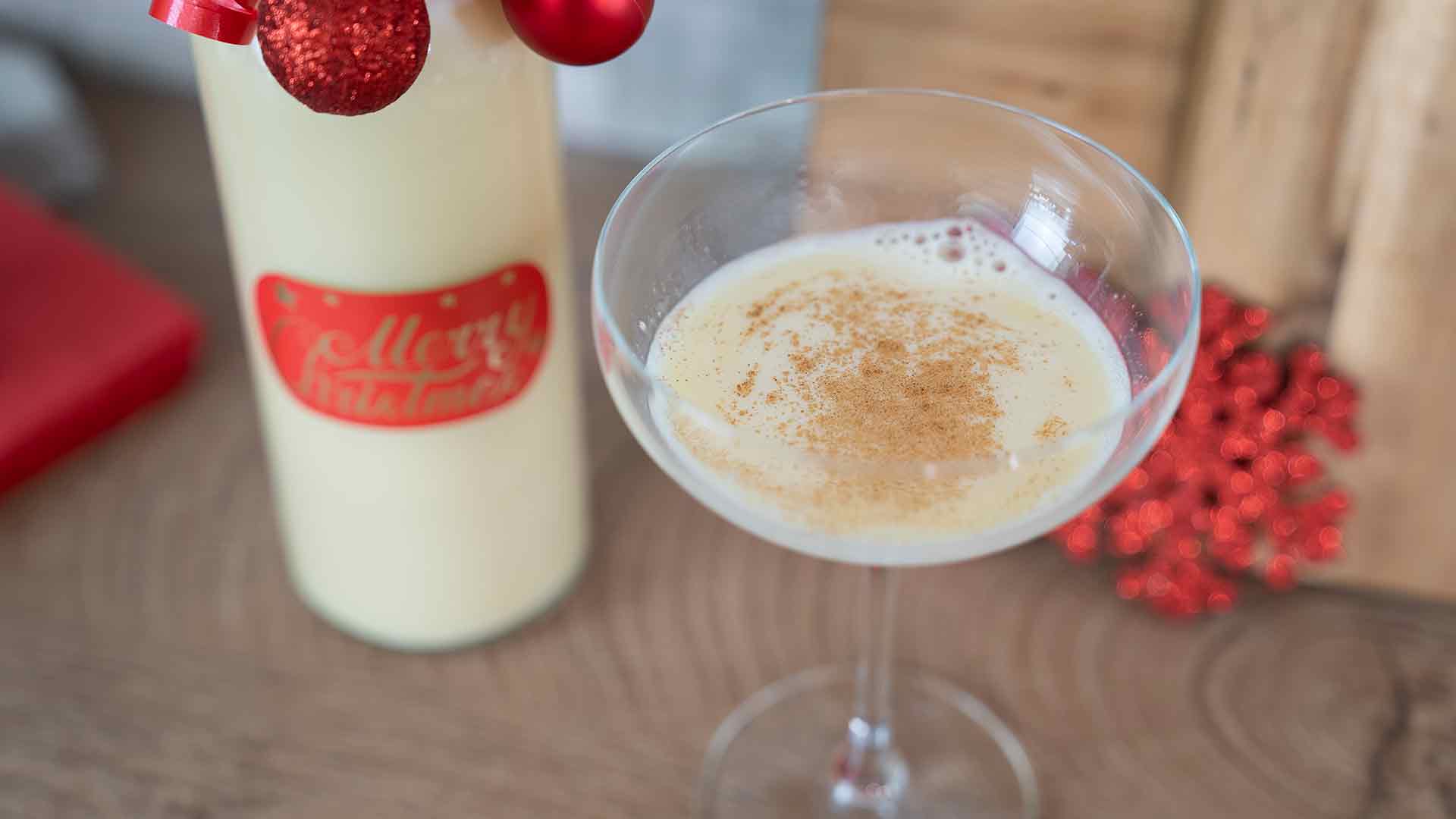 Ein Glas cremiger Eierlikör, bestreut mit Muskatnuss, steht neben einer festlichen Flasche, die mit roten Ornamenten und einem roten Etikett verziert ist, auf einer Holzoberfläche mit Weihnachtsdekor im Hintergrund.