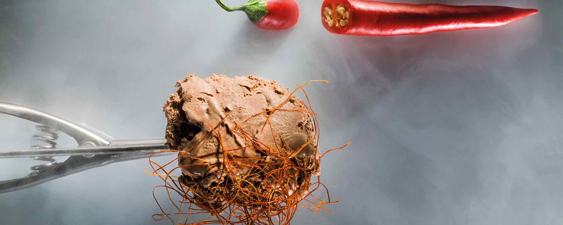 Ein Eisportionierer aus Metall hält eine Kugel Schokoladeneis mit dünnen Chilifäden. Darüber sind eine ganze rote Chilischote und eine geschnittene Chilischote auf einem rauchgrauen Hintergrund platziert.