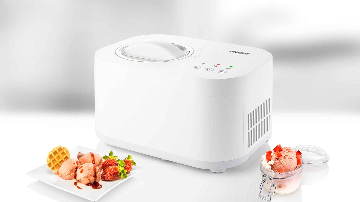 Eine weiße Eismaschine mit Knöpfen und Kontrollleuchten steht auf einer weißen Fläche. Daneben stehen Teller und ein Glas mit Kugeln buntem Eis, Erdbeeren und einer Waffel. Der Hintergrund ist unscharf.