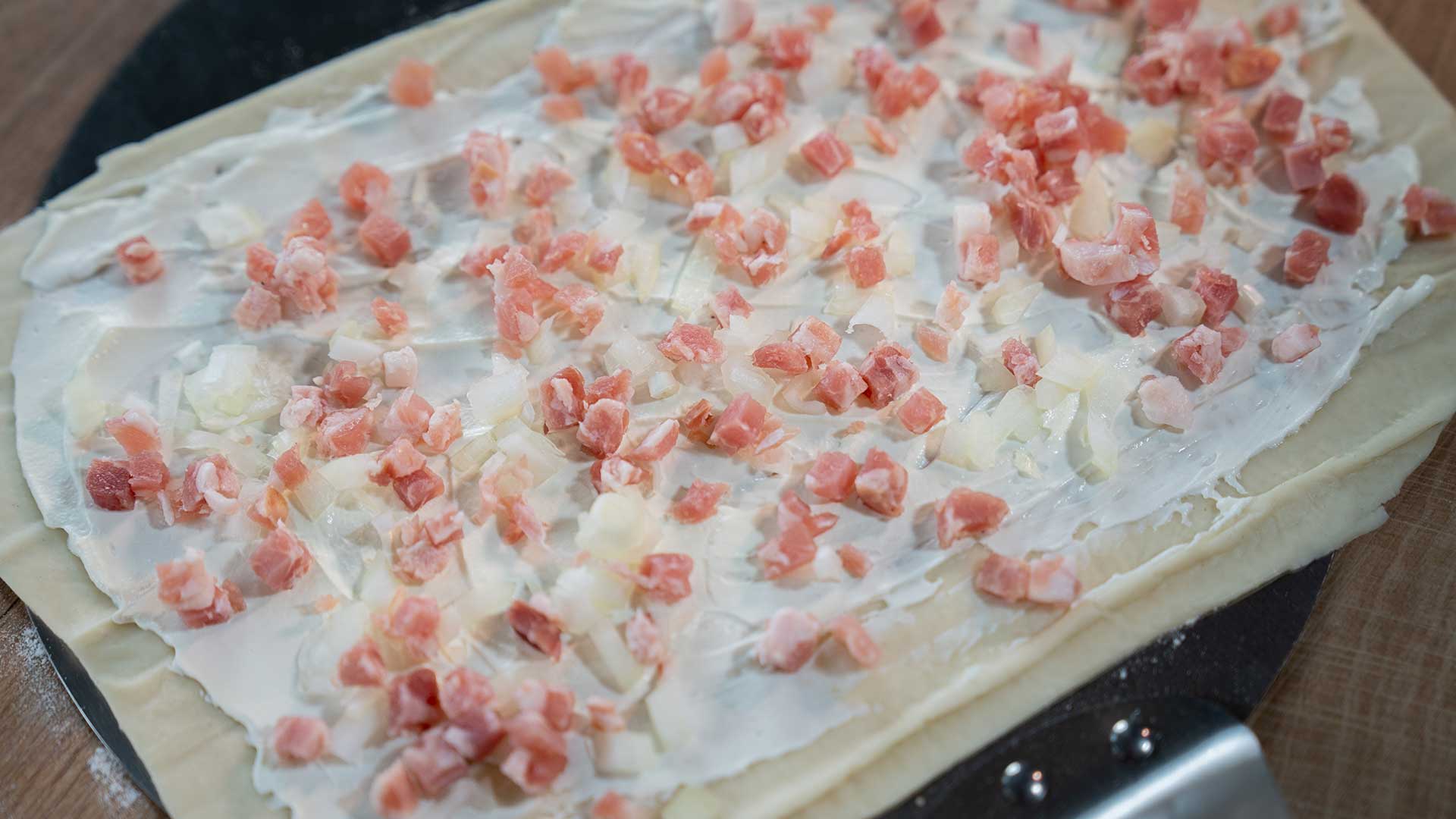Eine mit einer Schicht weißer Sahne bestrichene und mit kleinen Speck- und Zwiebelwürfeln belegte Teigplatte, zubereitet auf einer Holzoberfläche.