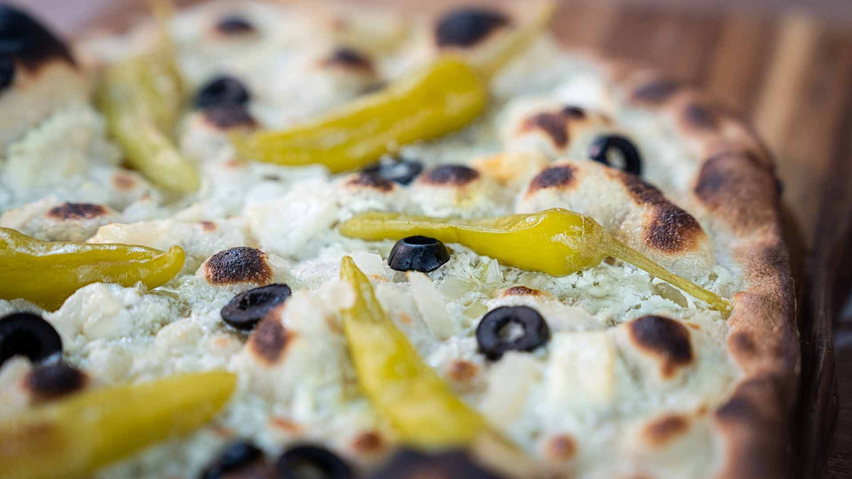 Eine Nahaufnahme einer Pizza mit Peperoncini-Paprika, schwarzen Oliven und geschmolzenem Käse auf einem goldenen, leicht verkohlten Rand, serviert auf einer Holzoberfläche.