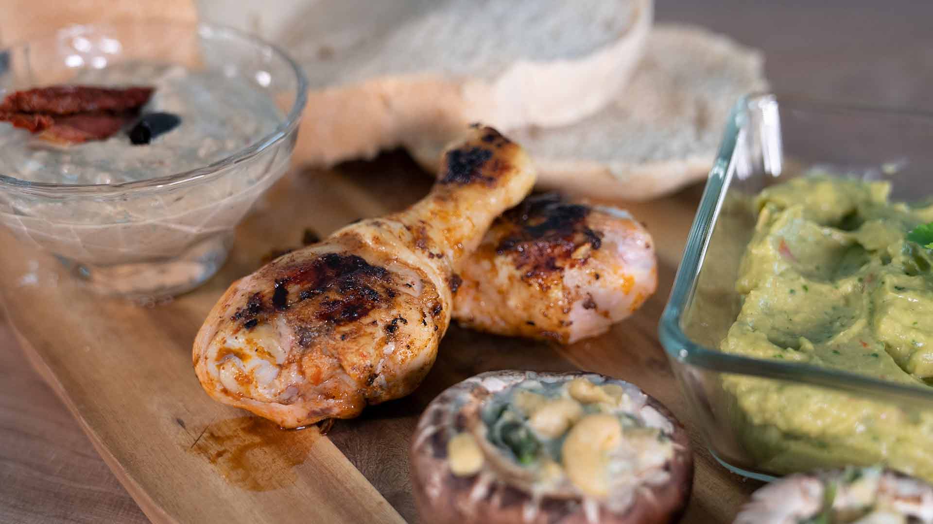 Zwei gegrillte Hähnchenkeulen auf einem Holzbrett, serviert mit geschnittenem Brot, einer Schüssel Guacamole, einer Schüssel Dip mit sonnengetrockneten Tomaten und einem gefüllten Champignon.