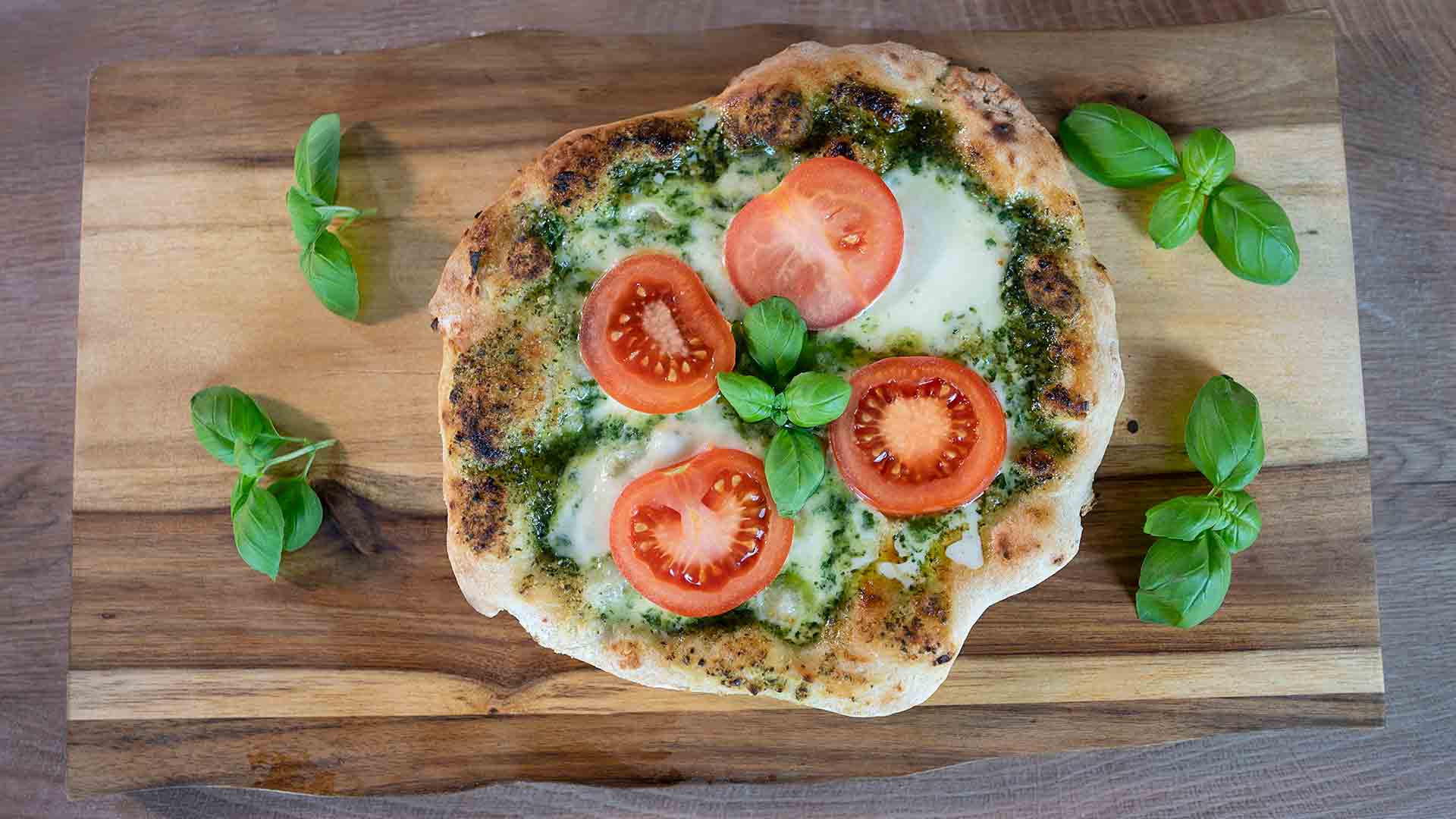 Eine rustikale Pizza mit geschmolzenem Käse, grünem Pesto und frischen Tomatenscheiben, garniert mit Basilikumblättern, liegt auf einem Holzschneidebrett.