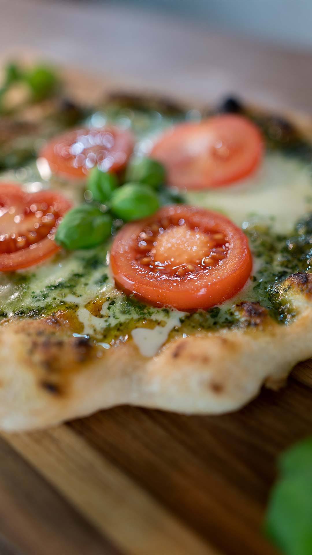 Nahaufnahme einer frisch gebackenen Pizza mit geschmolzenem Käse, Tomatenscheiben, grünem Pesto und frischen Basilikumblättern auf einem Holzbrett.