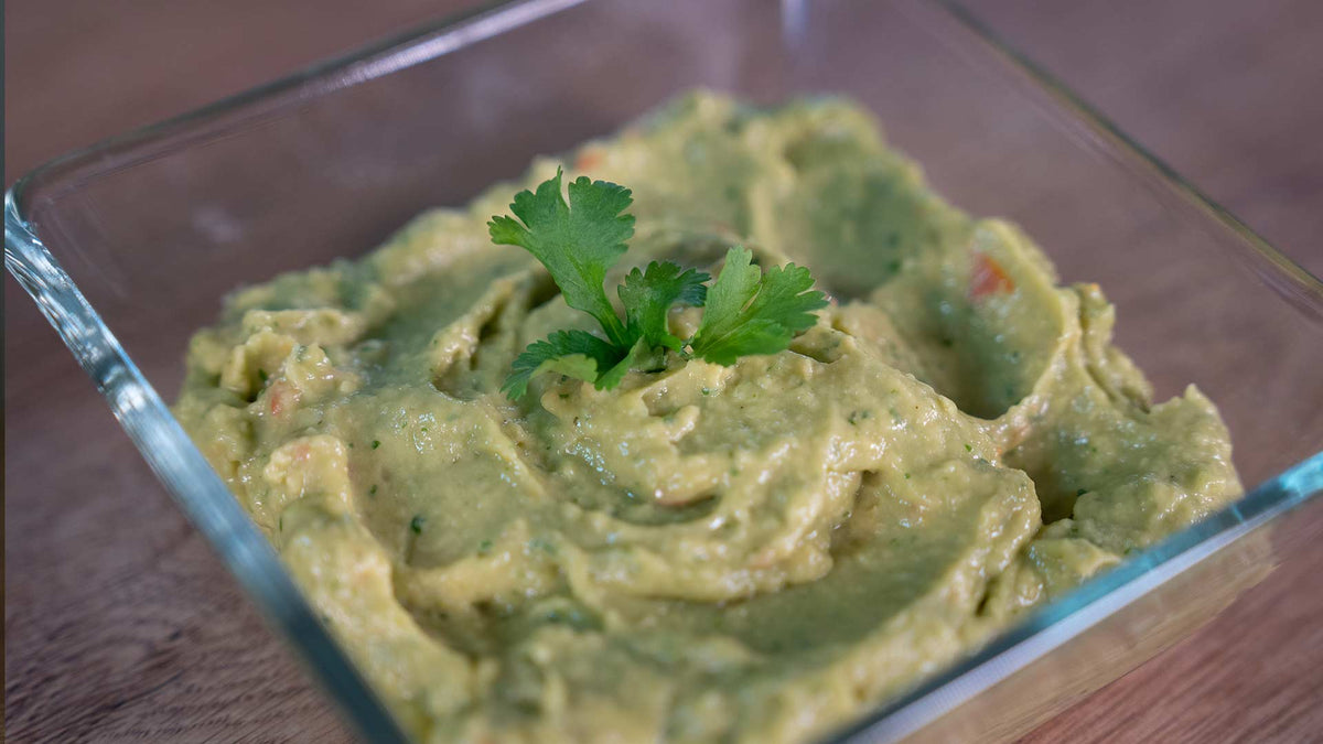 Auf einer Holzoberfläche steht eine durchsichtige Glasschale mit cremiger Guacamole und einem Zweig frischem Koriander.
