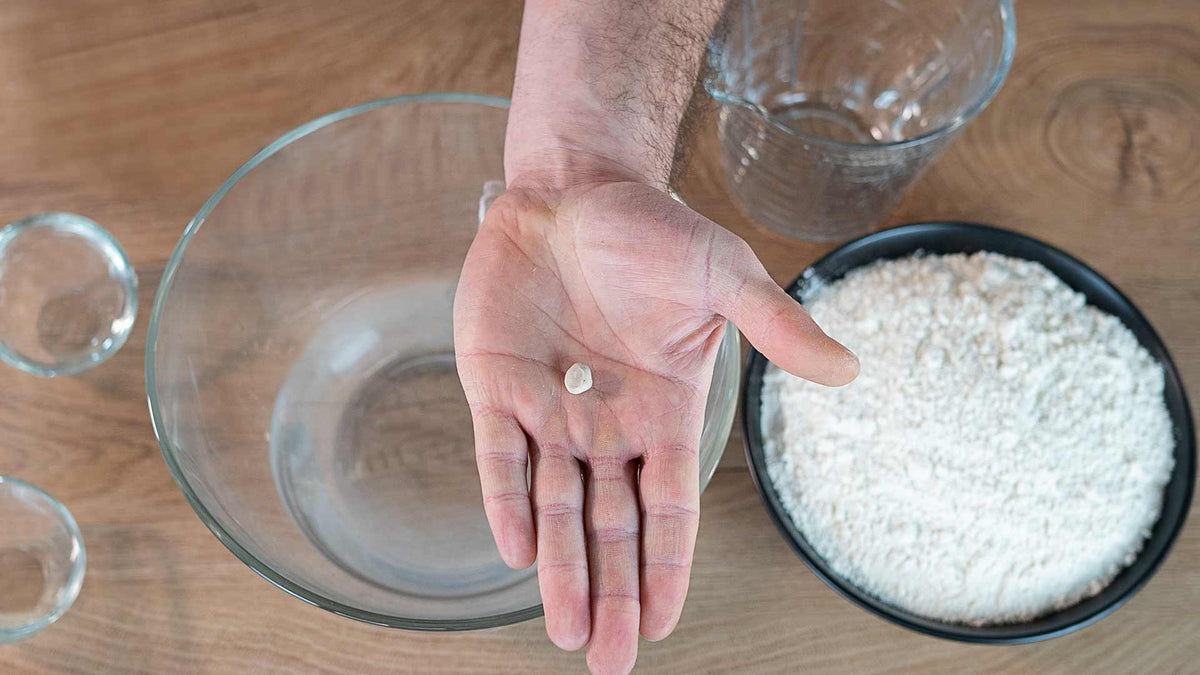 Eine Hand hält ein kleines Stück Hefe über einer durchsichtigen Rührschüssel, umgeben von Schüsseln mit Mehl und Wasser auf einer Holzoberfläche.