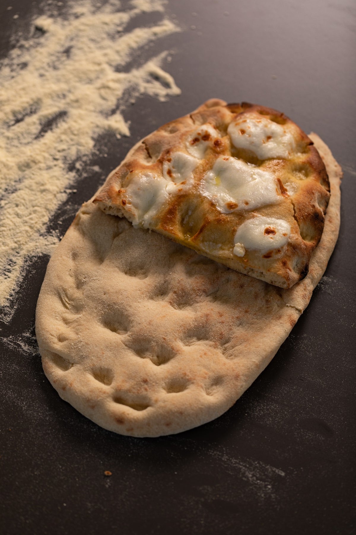 Ovales Fladenbrot auf dunkler Oberfläche, auf einer Hälfte mit geschmolzenem Käse belegt, daneben mit Mehl bestreut. Die andere Hälfte ist glatt und weist Einkerbungen vom Backen auf.