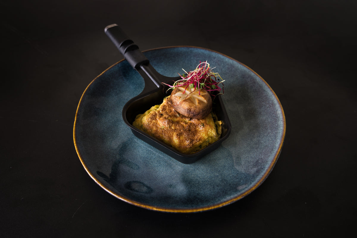 Genießen Sie ein Gourmetgericht, zubereitet mit dem Unold Raclette Smokeless inklusive Raclette Buch: ein goldbraunes Omelett mit Pilzen und Microgreens, elegant serviert in einer Pfanne auf einem blauen Teller.