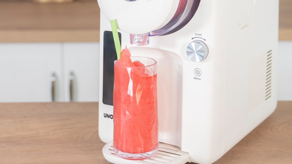 Eine Tischmaschine gibt leuchtend roten Slushy in ein klares Glas mit grünem Strohhalm aus, das auf einer Holzfläche in einer modernen Küche steht.