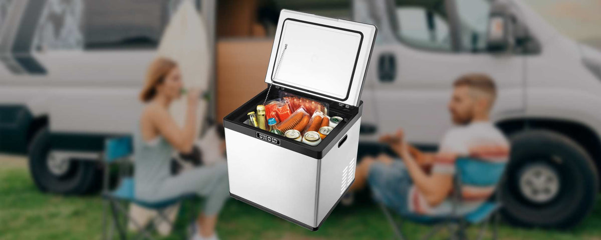 Ein tragbarer Kühlschrank mit Getränken und Lebensmitteln steht offen im Freien, im Hintergrund sitzen ein Mann und eine Frau auf Campingstühlen neben einem Lieferwagen.