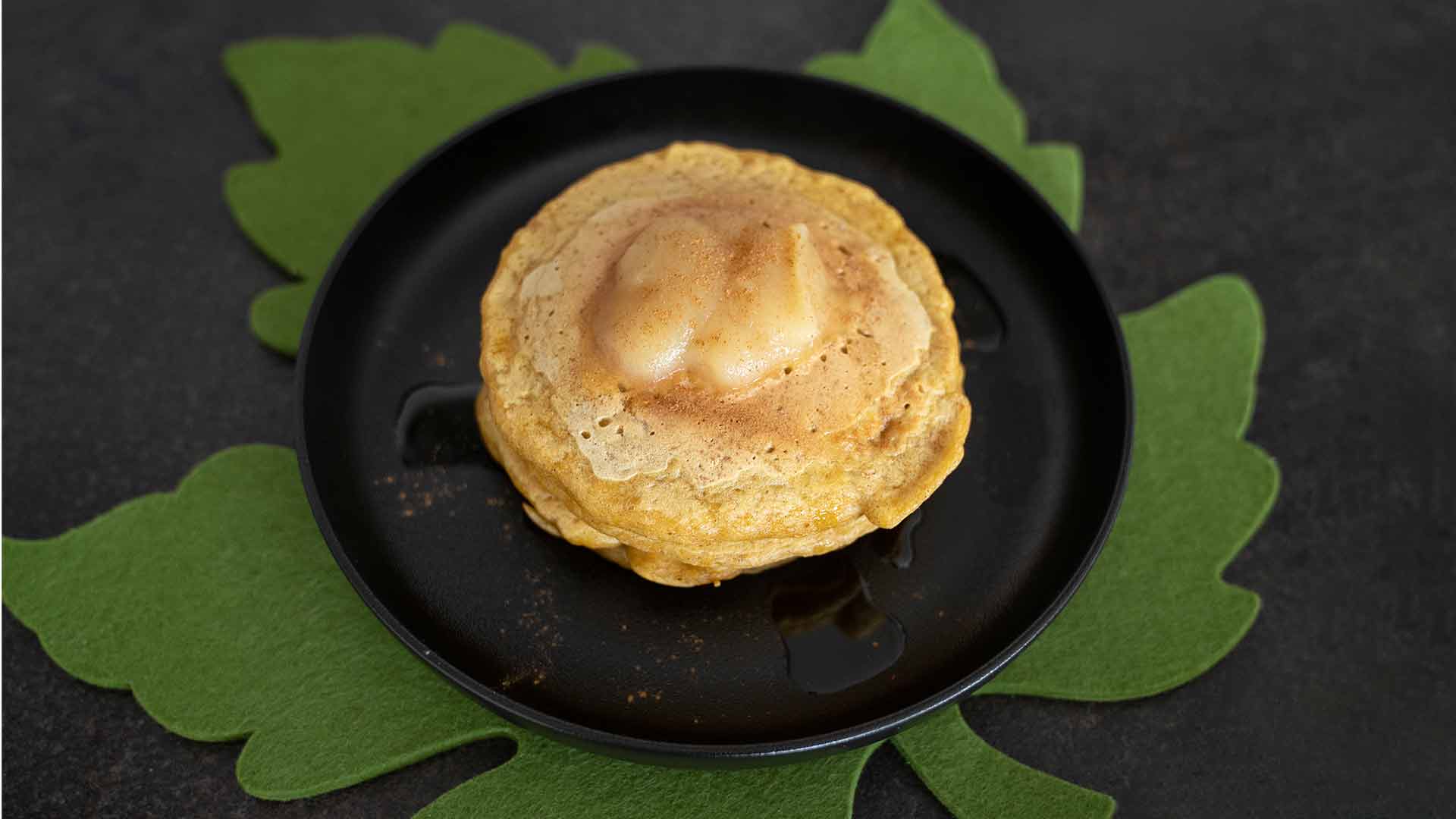 Ein Stapel Pfannkuchen mit einem Stück Butter und Sirup liegt auf einem schwarzen Teller mit einem grünen, blattförmigen Tischset darunter vor einem dunklen Hintergrund.