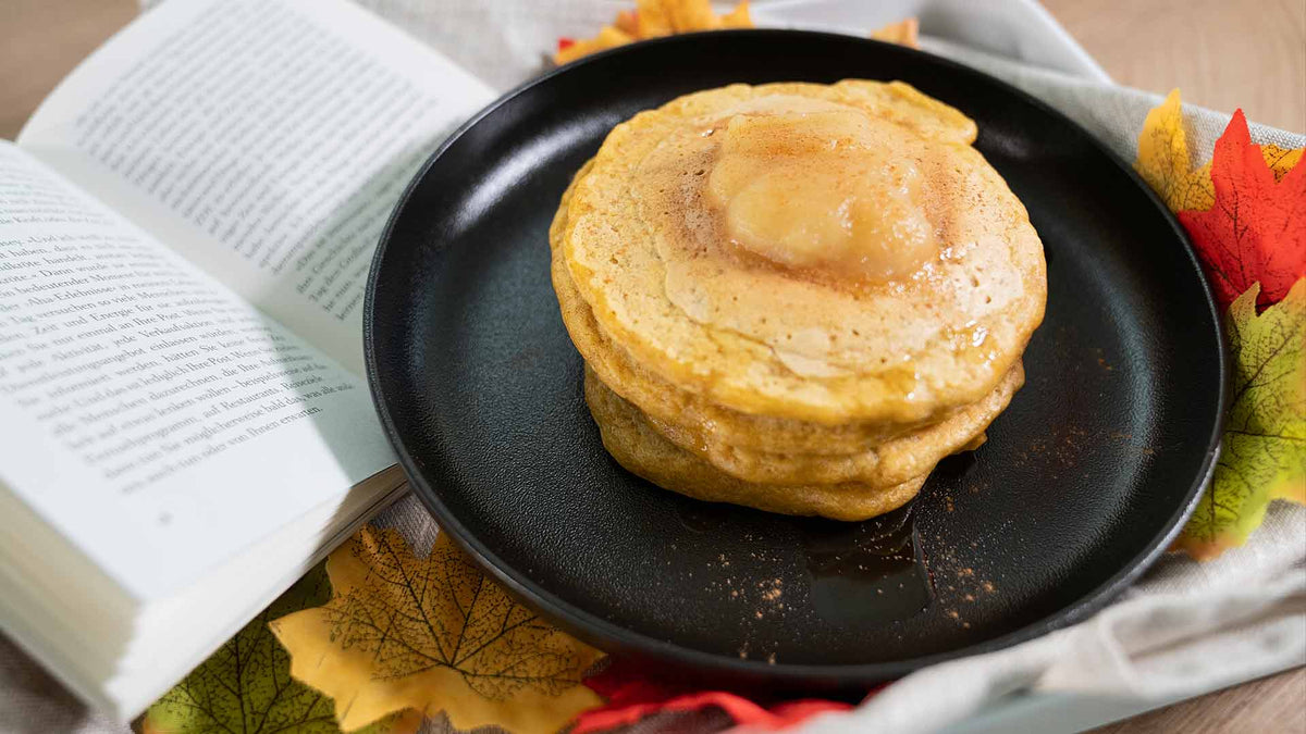 Ein Stapel Pfannkuchen mit geschmolzener Butter liegt auf einem schwarzen Teller, umgeben von buntem Herbstlaub. Daneben liegt ein offenes Buch und sorgt für eine gemütliche, weihnachtliche Atmosphäre.