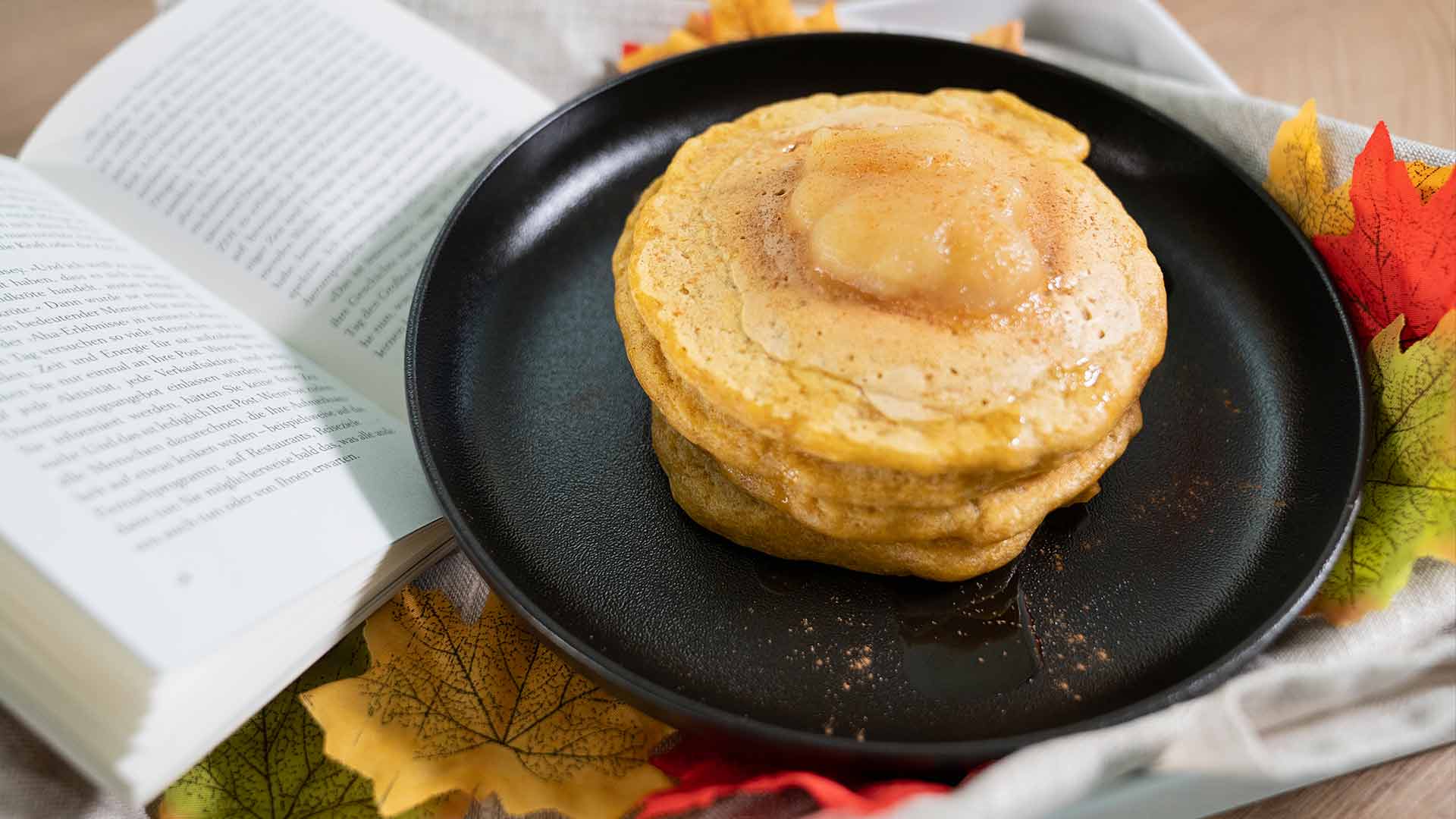Ein Stapel Pfannkuchen mit Butter darauf liegt auf einem schwarzen Teller, umgeben von Herbstblättern und einem offenen Buch, was eine gemütliche Herbstatmosphäre schafft.