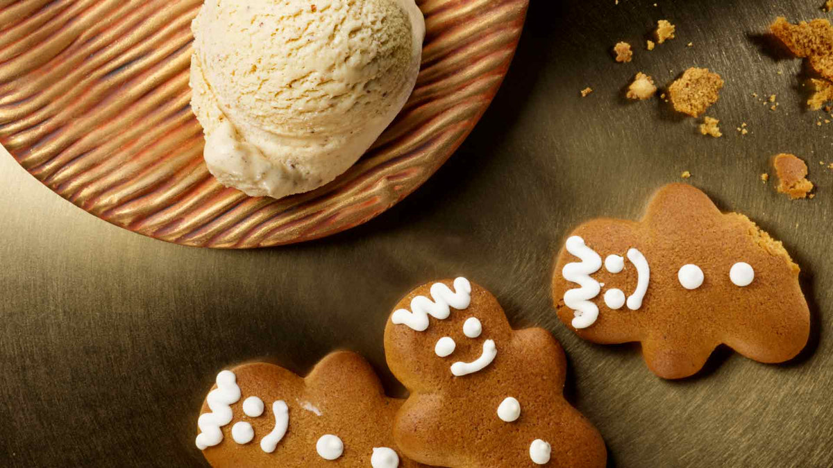 Drei Lebkuchen mit Gesichtern aus weißem Zuckerguss liegen auf einer goldenen Oberfläche neben einem braunen Teller mit einer Kugel Vanilleeis. In der Nähe sind Kekskrümel verstreut.