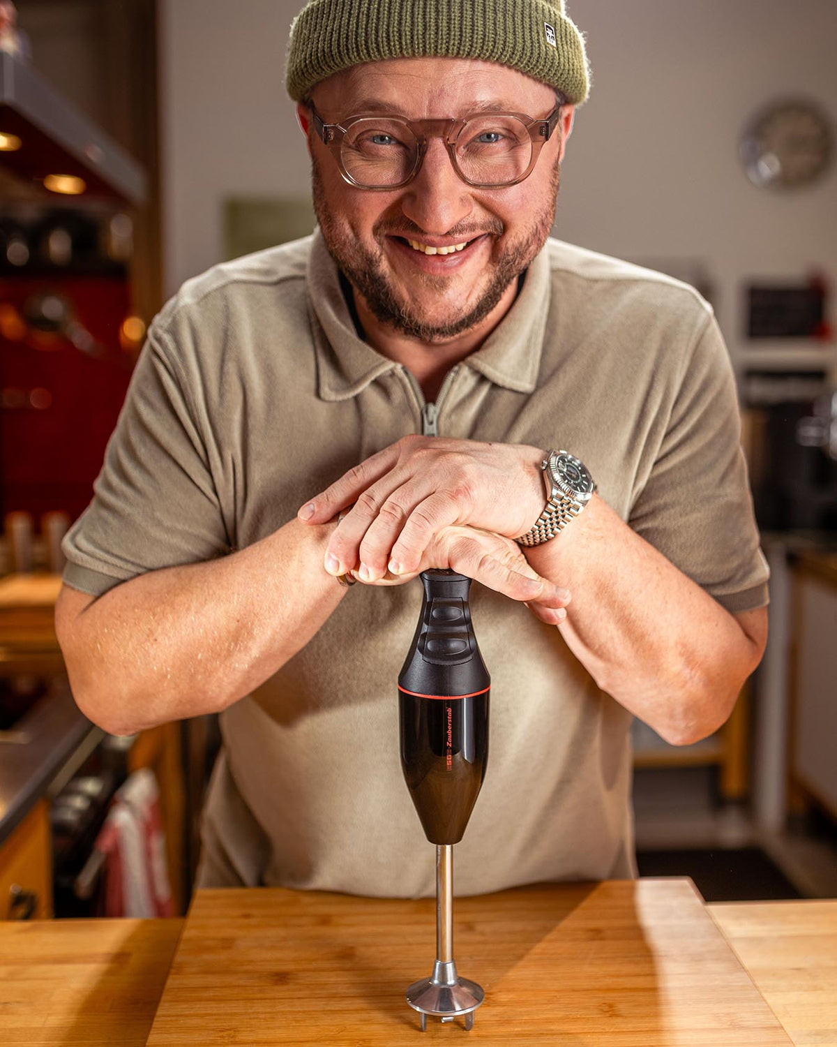 Ein lächelnder Mann mit Brille und grüner Mütze lehnt sich in einer gemütlichen Küche auf einen Stabmixer und legt seine Hände darauf.