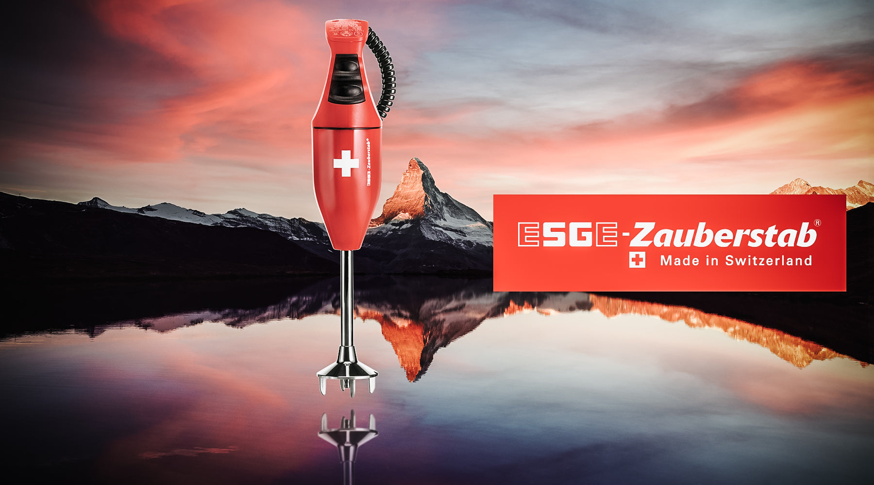 Vor einem dramatischen Sonnenuntergang über den Bergen und dem See steht ein roter ESGE-Zauberstab-Stabmixer mit einem weißen Schweizer Kreuz. Auf der rechten Seite befindet sich ein passendes rotes Logo mit der Aufschrift „ESGE-Zauberstab Made in Switzerland“.