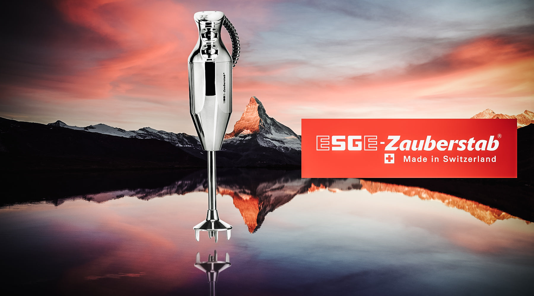 Ein ESGE-Zauberstab-Stabmixer mit silbernem Griff steht aufrecht auf einer reflektierenden Oberfläche, im Hintergrund ein Berg und ein farbenfroher Sonnenuntergangshimmel. Rechts sind das rote Logo der Marke und „Made in Switzerland“ zu sehen.